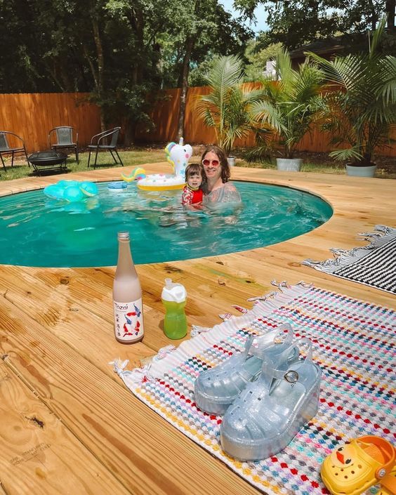 An in-ground stock tank pool with a wooden deck, potted plants and trees and some outdoor furniture.