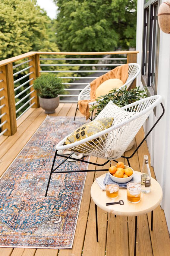 An inviting summer balcony with a boho rug, some round chairs, a table and potted greenery plus pillows.