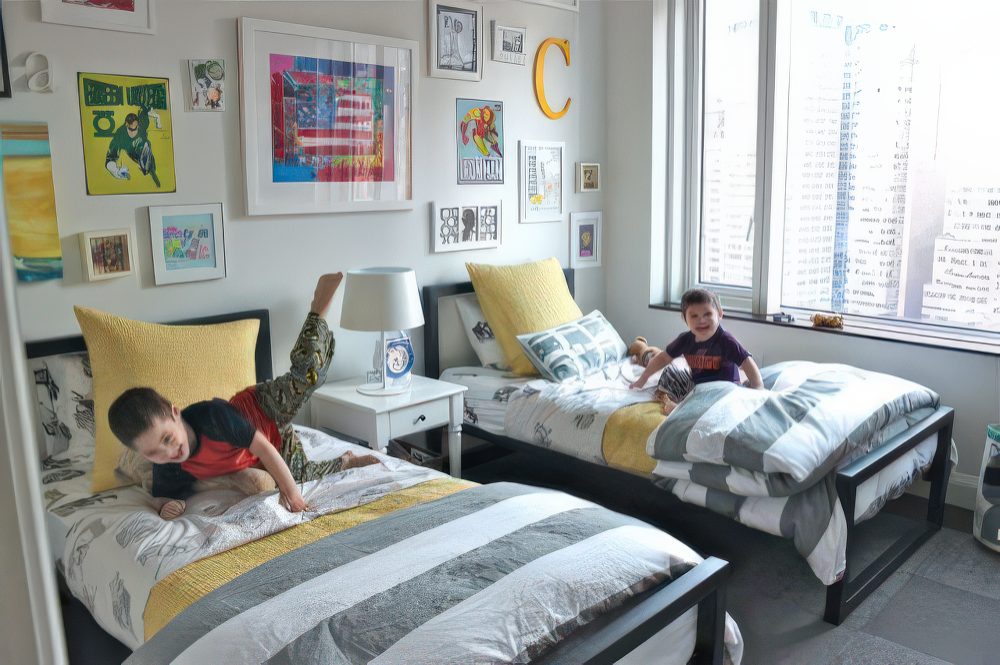 A contemporary twin bedroom featuring a mix of bright artwork, yellow pillows, and sleek black bed frames. A city skyline view through the window adds an urban vibe.