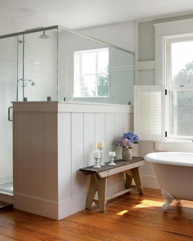 A neutral farmhouse bathroom with planked walls and a shower space, with a pony wall and a vintage bathtub plus a bench.