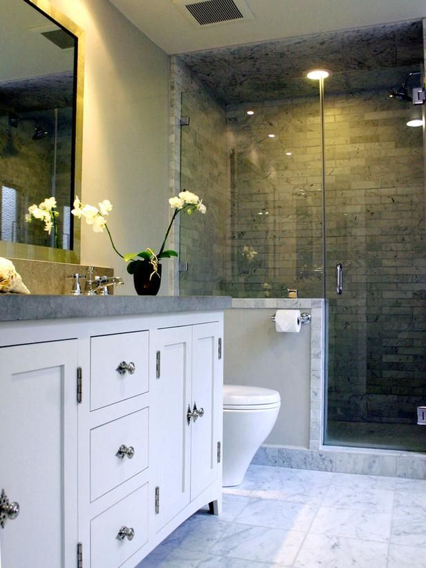 A chic bathroom with white walls, a shower space with a pony wall and grey tiles, a large white vanity with a grey stone countertop.