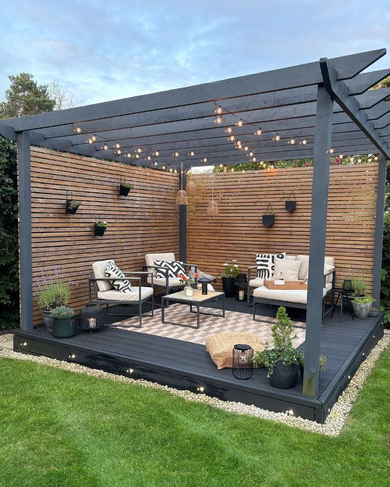 The dark raised deck is paired with a pergola with screens and lights; modern furniture and plants create a nice outdoor living room.