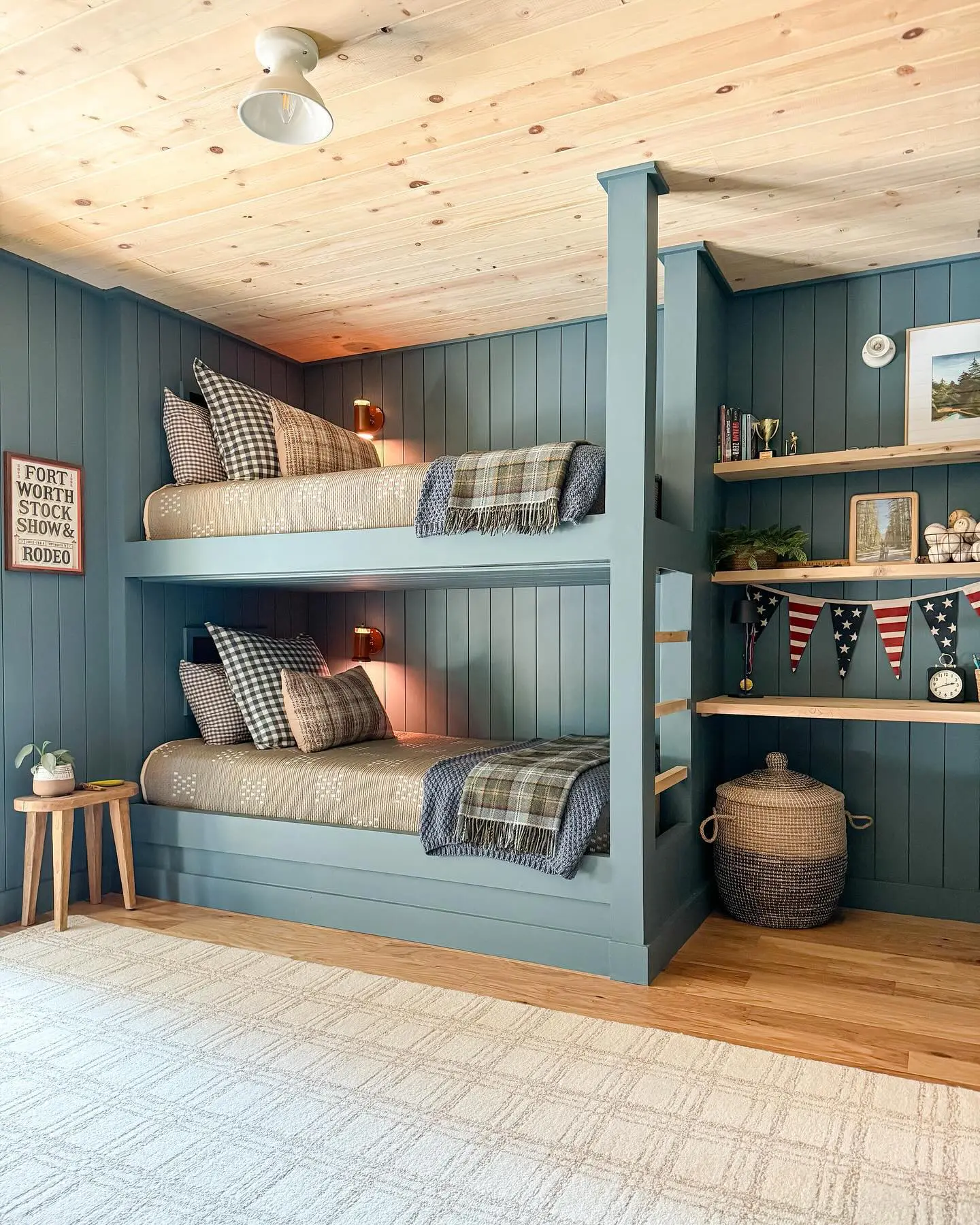 A warm and inviting bunk room with built-in blue bunk beds, wooden wall paneling, and cozy plaid bedding. Small wall sconces add a soft glow, while natural wood shelves display books and trophies.

(@girlandgrey).