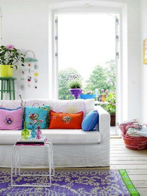 A bright and welcoming summer living room with bold floral pillows, with a colorful printed rug and some bold blooms in pots inside and outside, on the balcony.