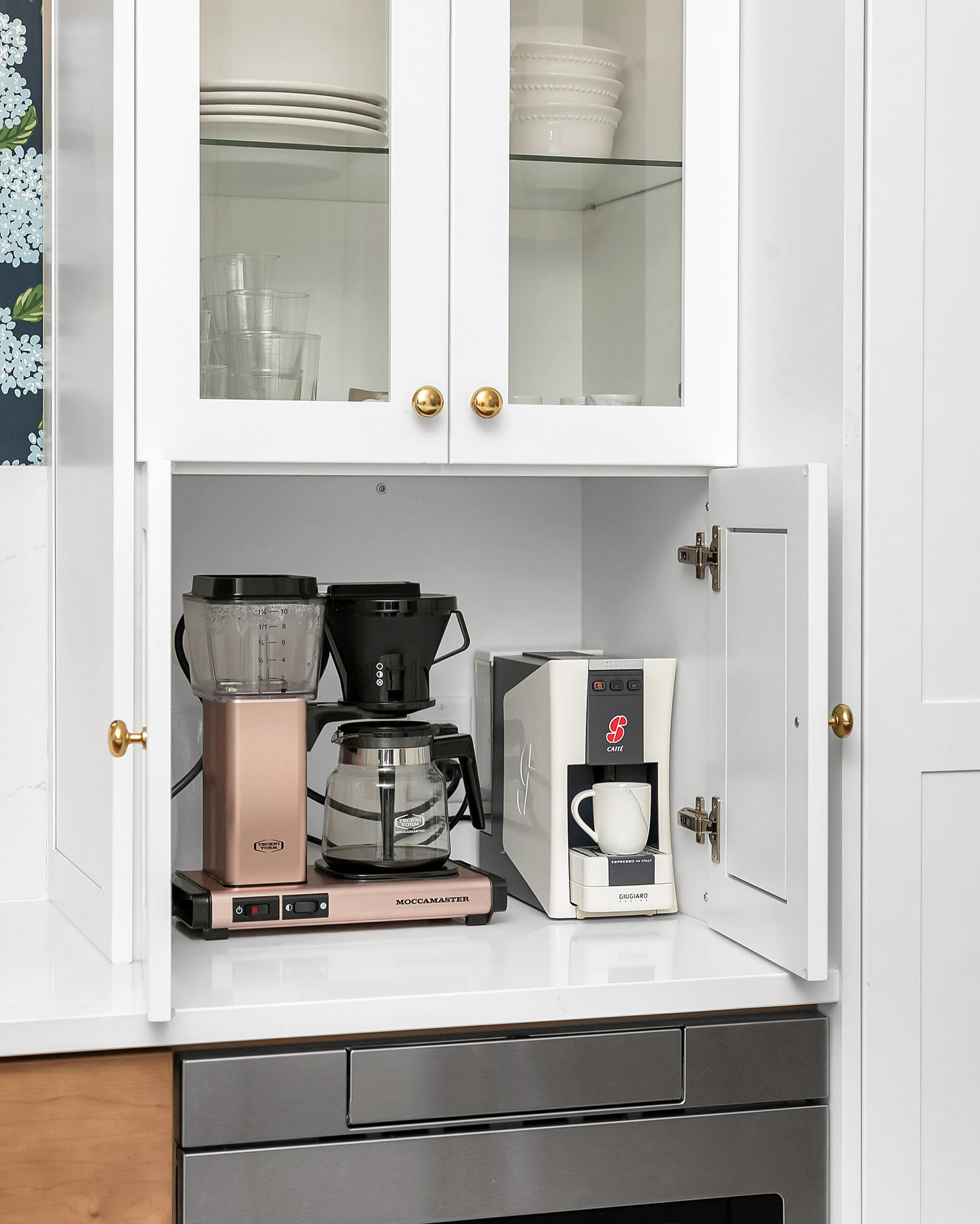 This smartly designed kitchen cabinet hides a coffee station behind elegant white doors with brass knobs. It neatly accommodates a Moccamaster coffee machine and a compact espresso maker.

(@designlinekitchens).