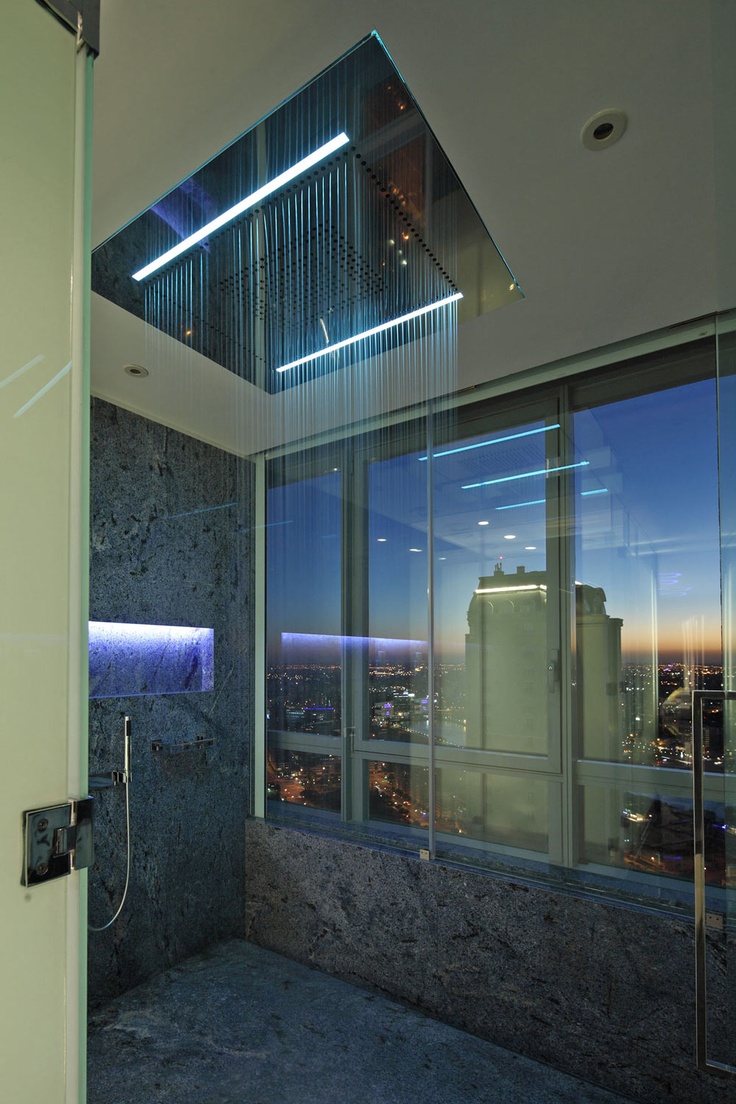 A gorgeous modern shower space with a view of the big city through a clear glass wall and built-in lights and a large rain shower.