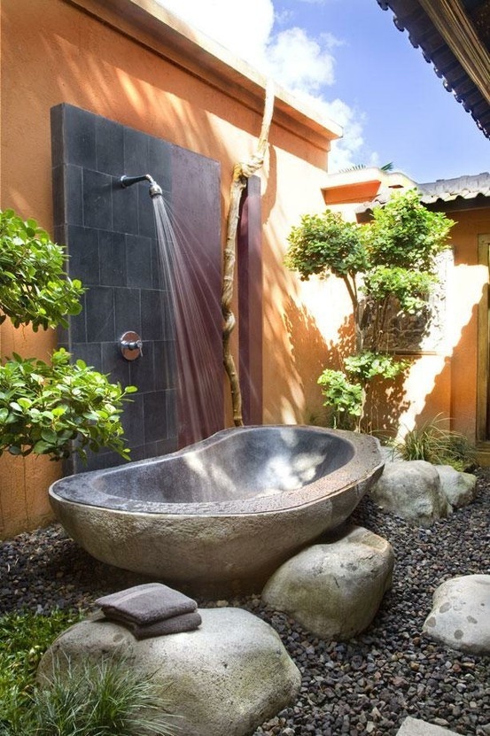 An outdoor tropical bathroom with a metal tub surrounded with rocks and a shower right here plus beautiful mini trees around.