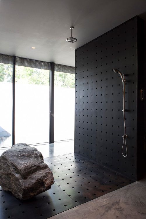A contemporary and dramatic bathroom with a black metal wall and floor, a large rock and a glazed wall to enjoy the views.
