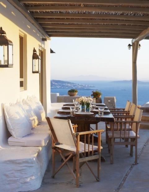A Mediterranean summer terrace with a built-in bench, wooden furniture, candle lanterns and blooms and greenery plus a cool sea view.