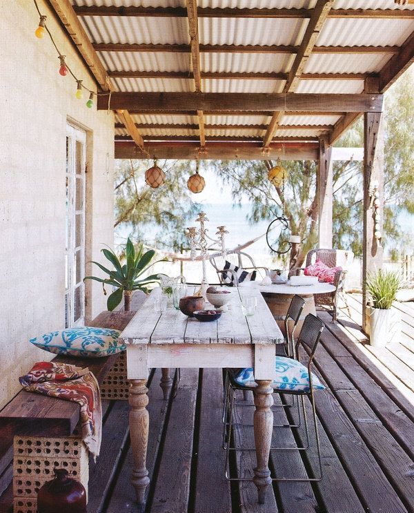 A bright tropical summer terrace with a whitewashed table, a carved wooden bench, metal chairs and textiles and lanterns attached to the roof.