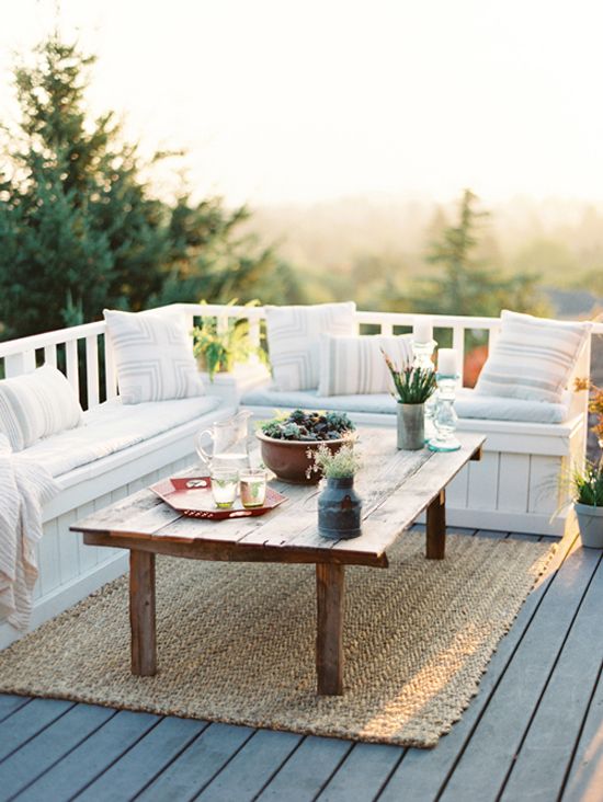 A simple and neutral summer terrace with a built-in storage bench, a wooden table, a jute rug, greenery and herbs in pots.