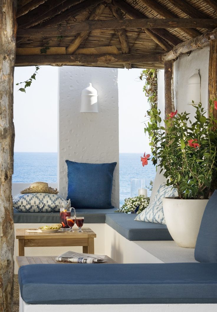 A seaside summer terrace in white, with blue textiles and upholstery, potted blooms and greenery and a cool seaside view.