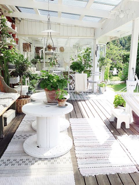 A white farmhouse terrace done with vintage rustic furniture in white, white textiles and lots of greenery in pots here and there.
