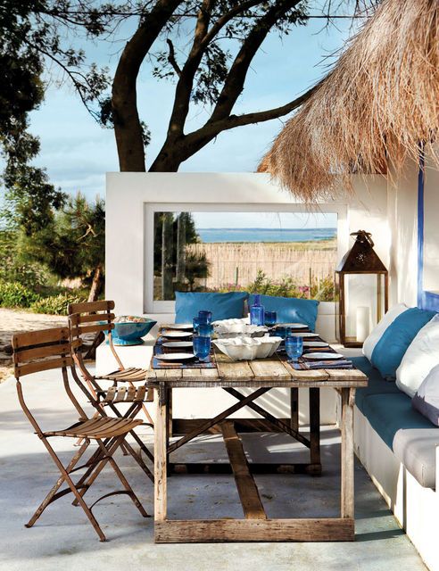 A bright summer seaside terrace with a built-in bench, wooden furniture, white and navy textiles, statement candle lanterns.