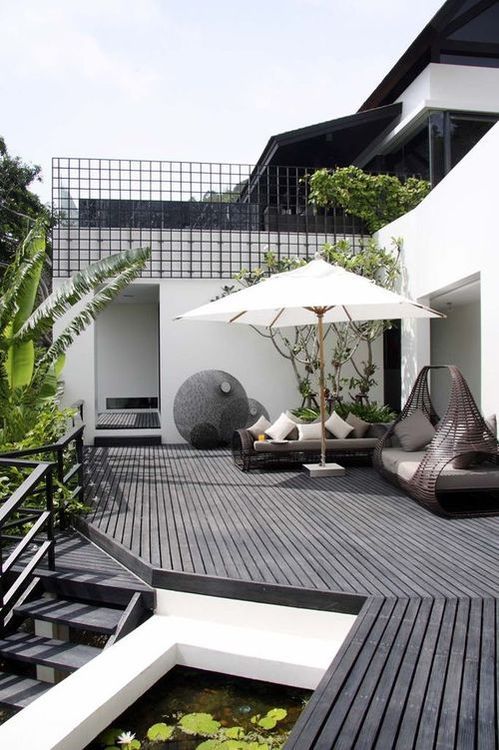 A monochromatic Scandinavian terrace with a black deck, dark rattan furniture, lots of greenery and a tree plus a little pond.
