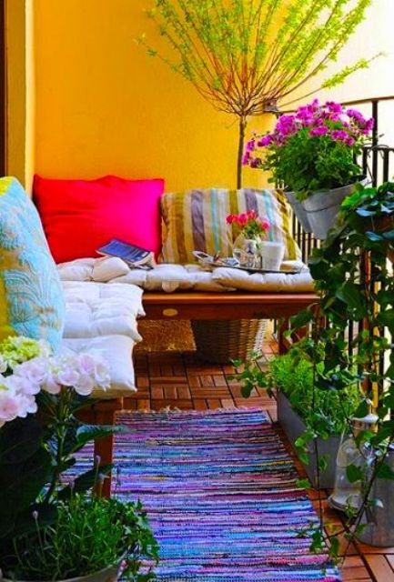 A bright and colorful summer balcony with yellow walls, benches with bright pillows, rugs and potted greenery and blooms.