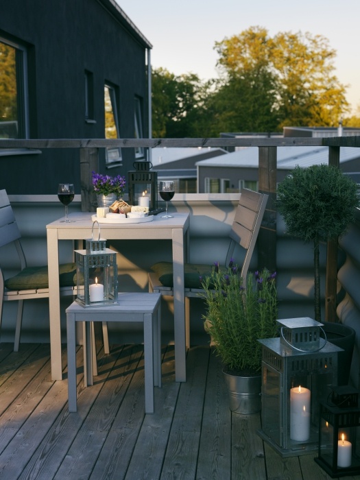 A small and cozy summer balcony with grey wooden furniture, potted greenery and blooms and candle lanterns.