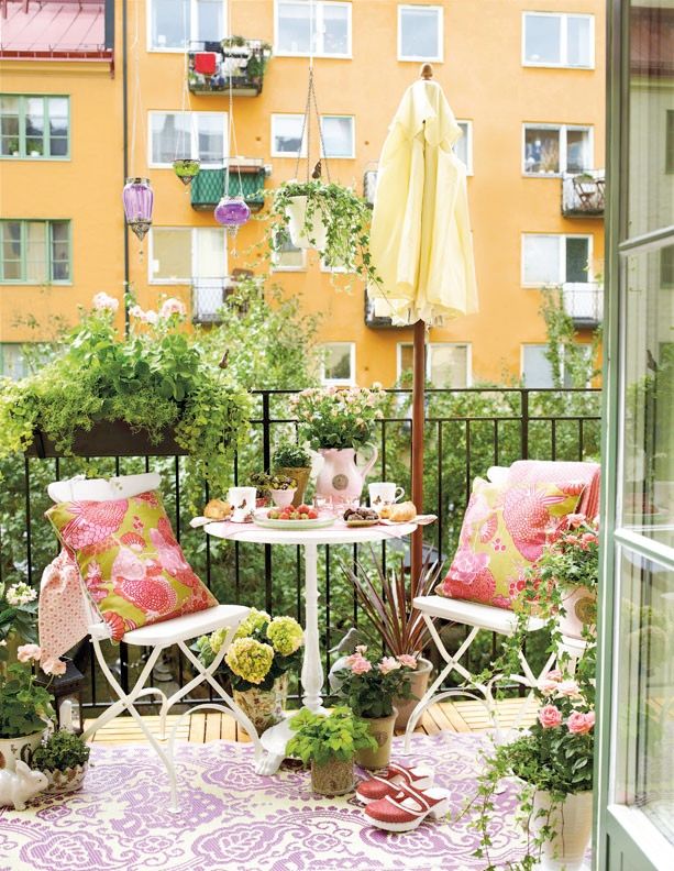 A welcoming summer balcony with simple white metal furniture, colorful printed textiles, potted blooms and greenery.