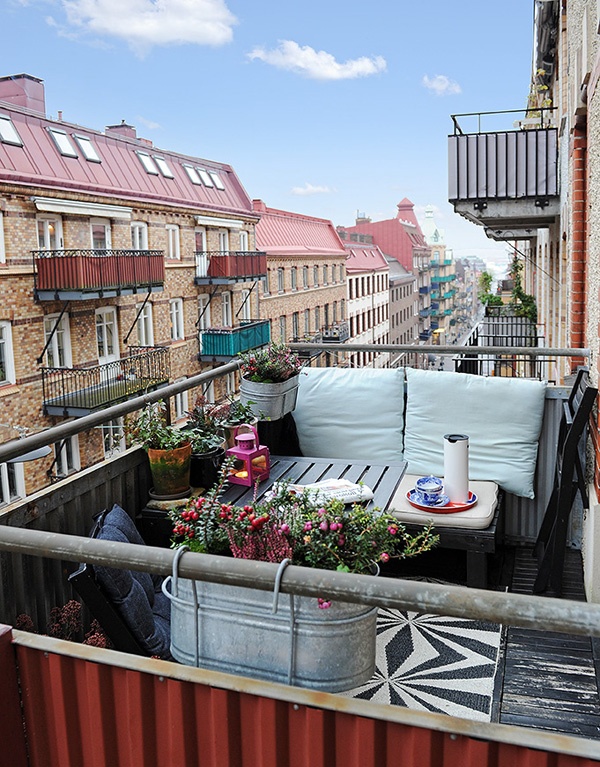 A small summer balcony with wooden furniture, neutral textiles, a pink candle lantern and potted greenery and blooms.