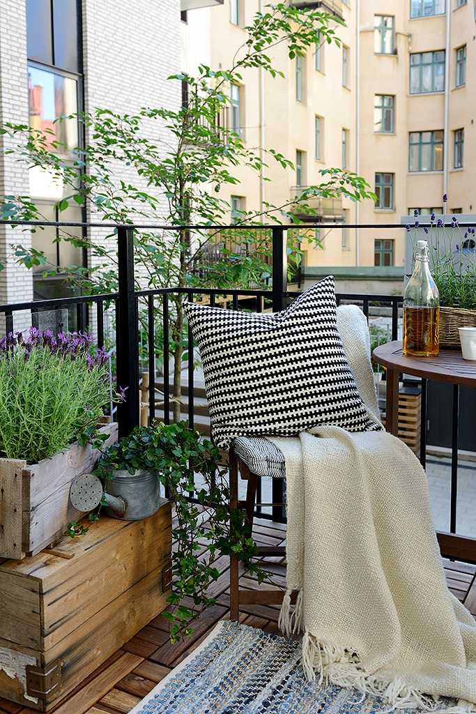 A welcoming summer balcony with wooden crates with greenery and blooms, folding wooden furniture and neutral textiles.