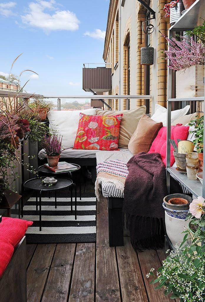 A bright summer balcony with a dark wooden floor, dark wooden furniture, potted greenery and blooms and bright textiles.