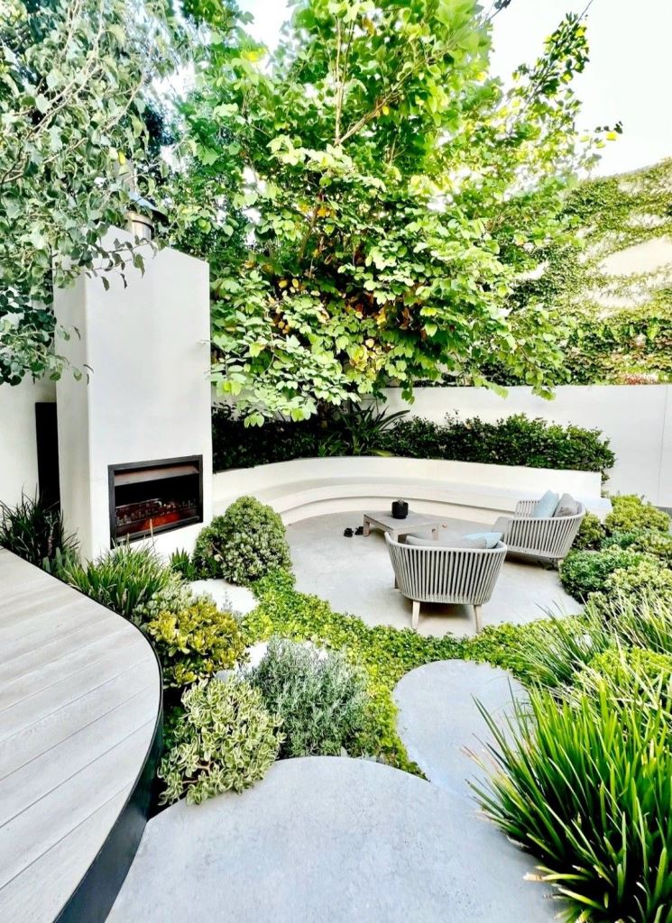 The refined space is composed of a raised garden bed with greenery and a built-in bench, a fireplace and some seating furniture.