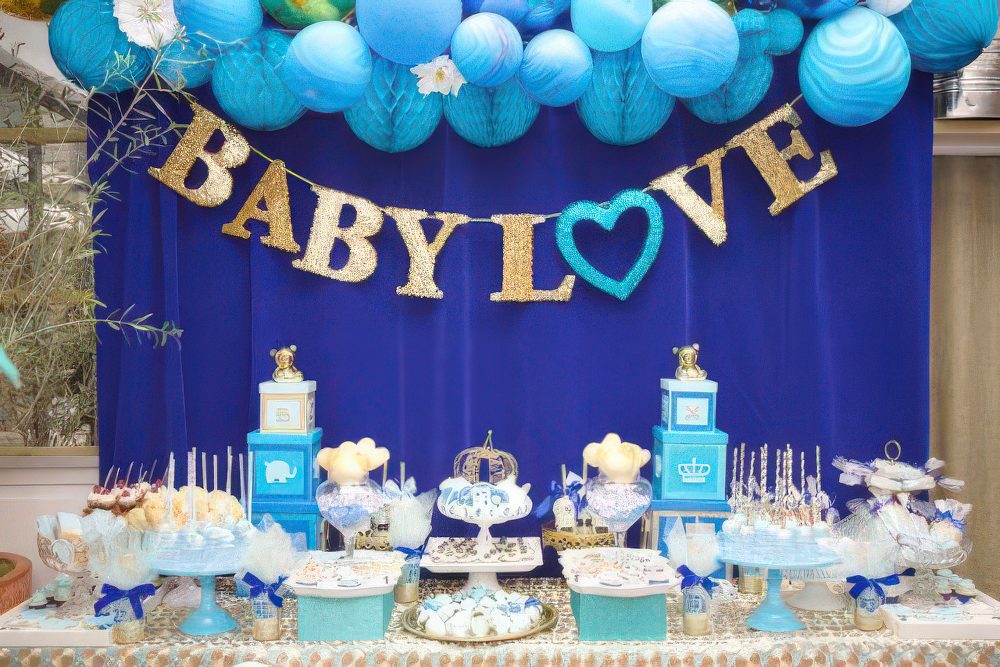 A bold blue and gold dessert table with blue balloons and pompoms, a letter banner and bright blue and gold stands.