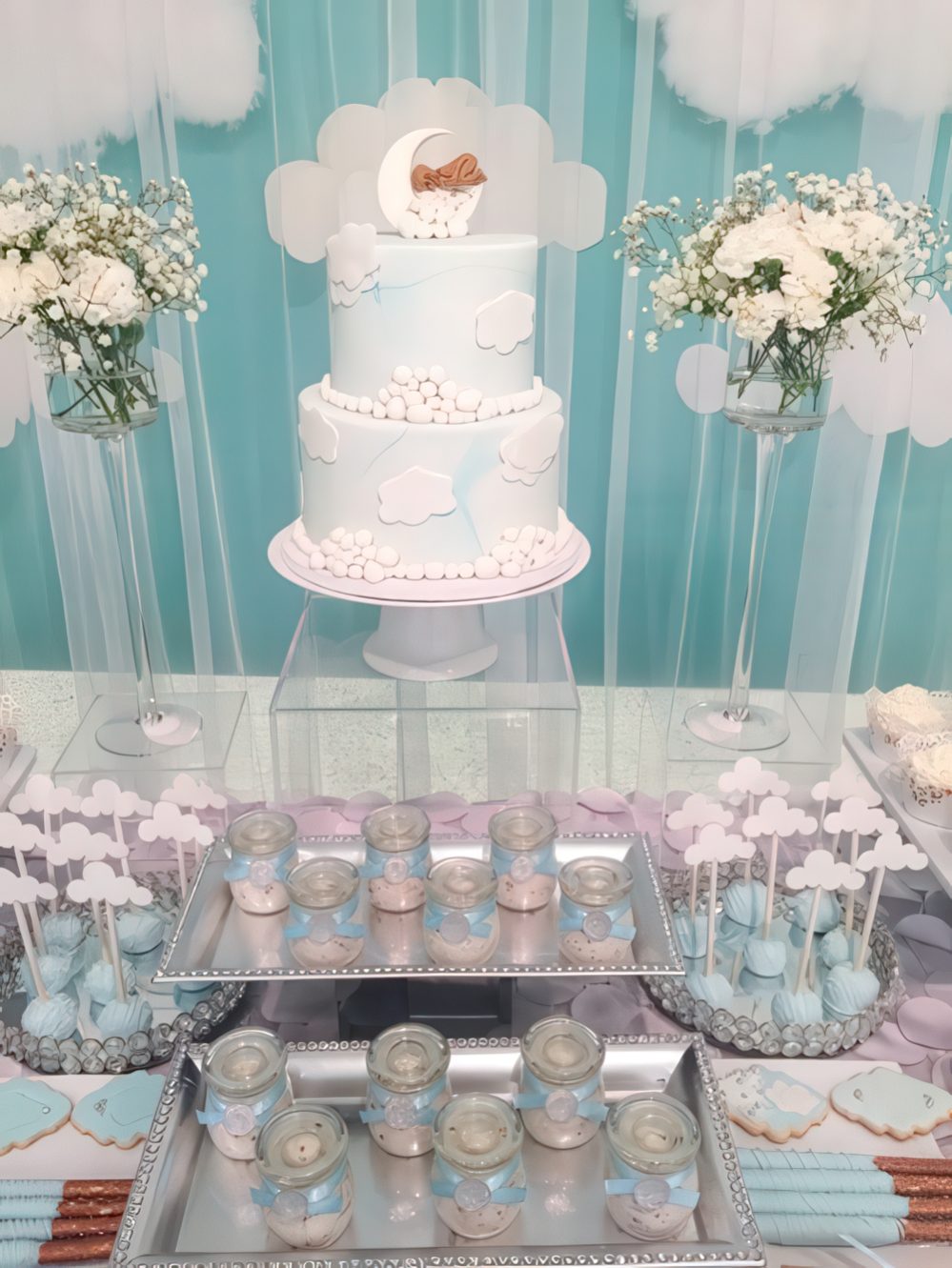 A blue and white dessert table with white floral arrangements, fake clouds, blue and white desserts and a cake.