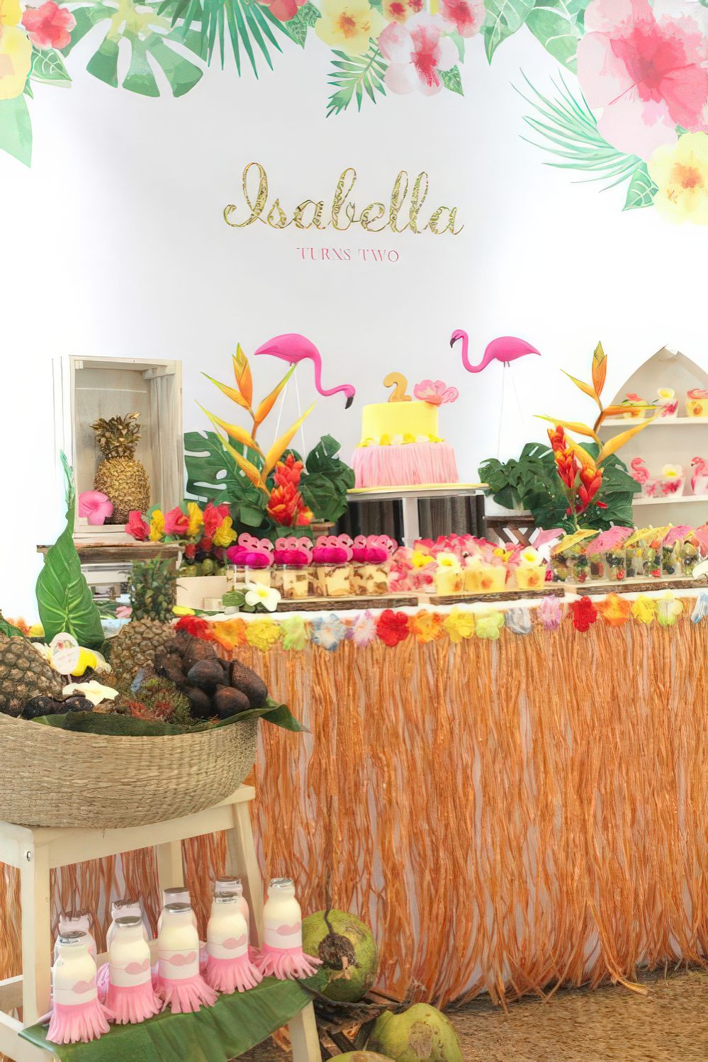 A tropical dessert table with touches of yellow, pink, green, orange, flamingos and fruits in a basket.