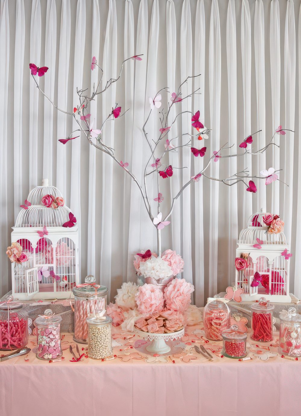 A pink and white baby shower table with vintage cages, a whitewashed tree with pink butterflies, vintage stands with various sweets.