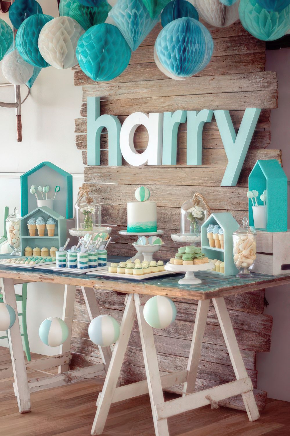 A turquoise and white baby shower dessert table with paper pompoms, house-shaped shelves and a trestle table.