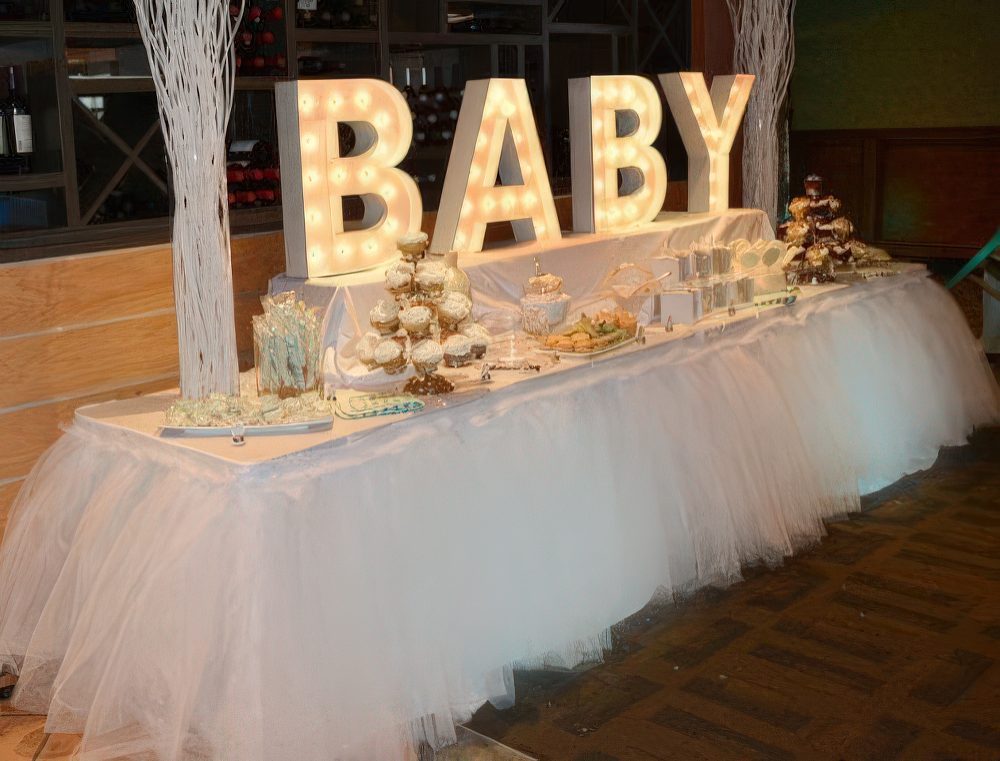 A bright white baby shower dessert table with lights, branch arrangements, a tulle tablecloth and lots of desserts.