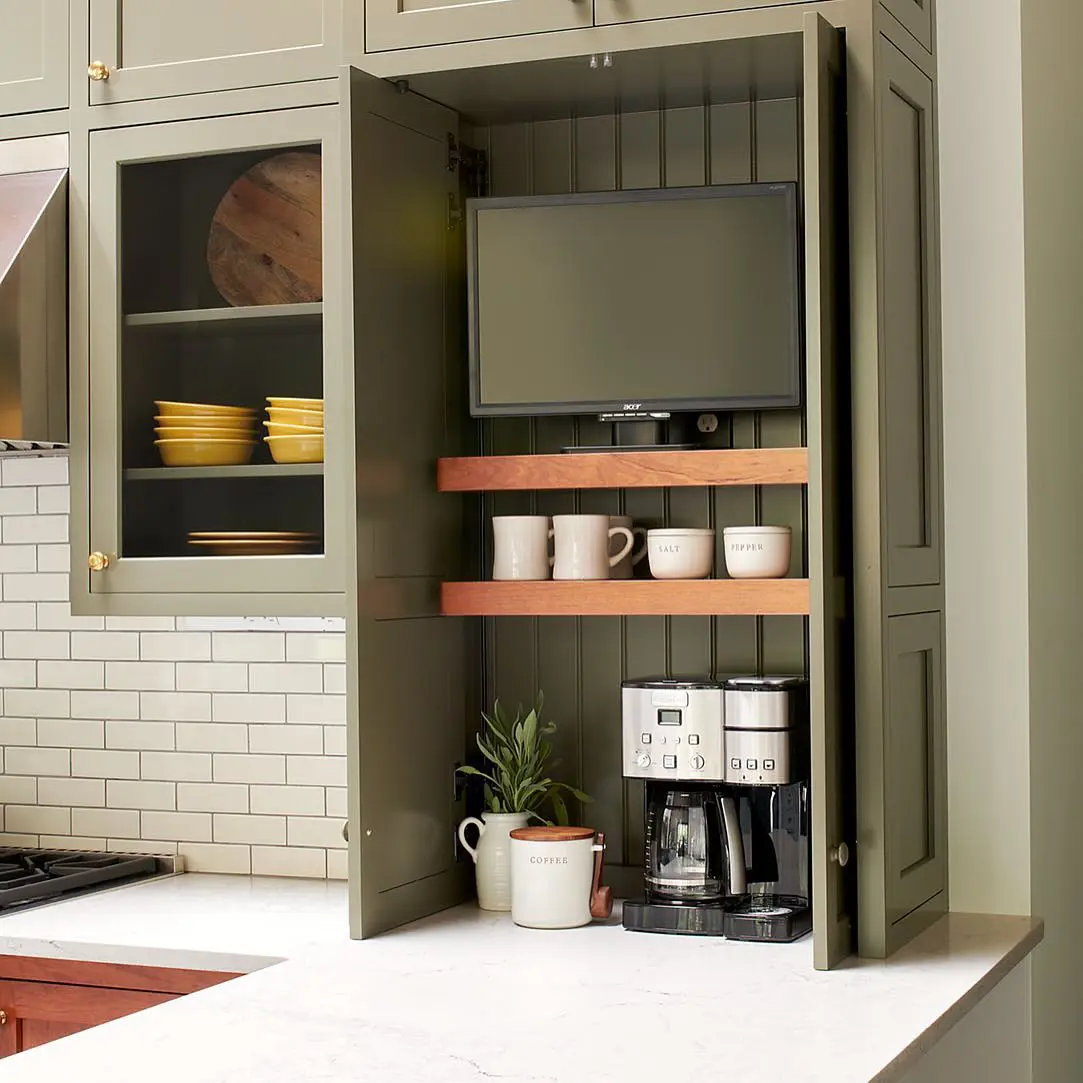 A unique coffee station inside a deep olive green cabinet features open shelving with wooden accents, providing a perfect spot for mugs, coffee beans, and accessories. The built-in TV adds a functional touch, making morning coffee routines more enjoyable.

(@airy_kitchens).