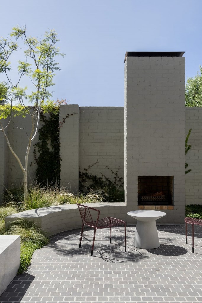The stylish modern patio is done with a brick fireplace and curved benches, with greenery and trees.