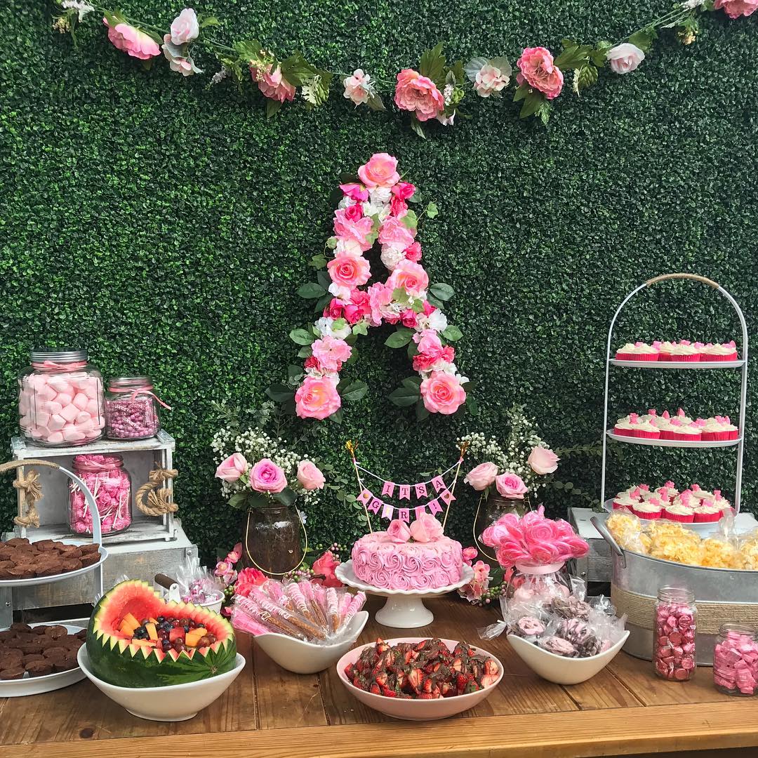 A pink-filled garden party dream with an oversized floral "A", lush greenery backdrop, and rows of sweet treats like rose cupcakes, chocolate-covered strawberries, and jars of pink candies.

(@kseventdecorations1).