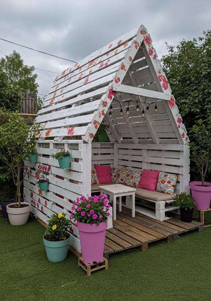 This house-shaped pergola features a sofa, also built of palelts, and lights on top. Bright blooms and painted flowers make the space cuter.