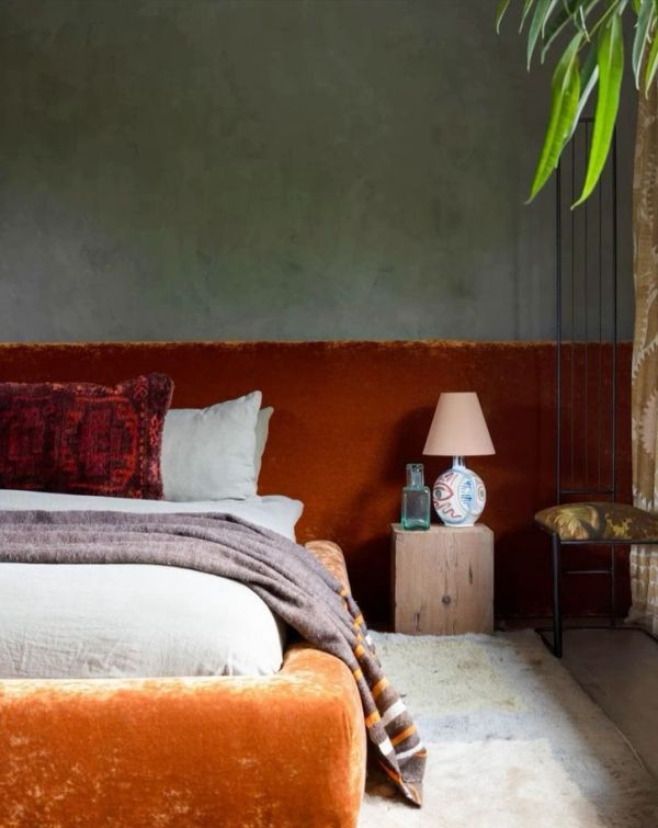 The moody bedroom is done with grey limewashed walls, an orange velvet upholstered bed with a matching wall to wall headboard.