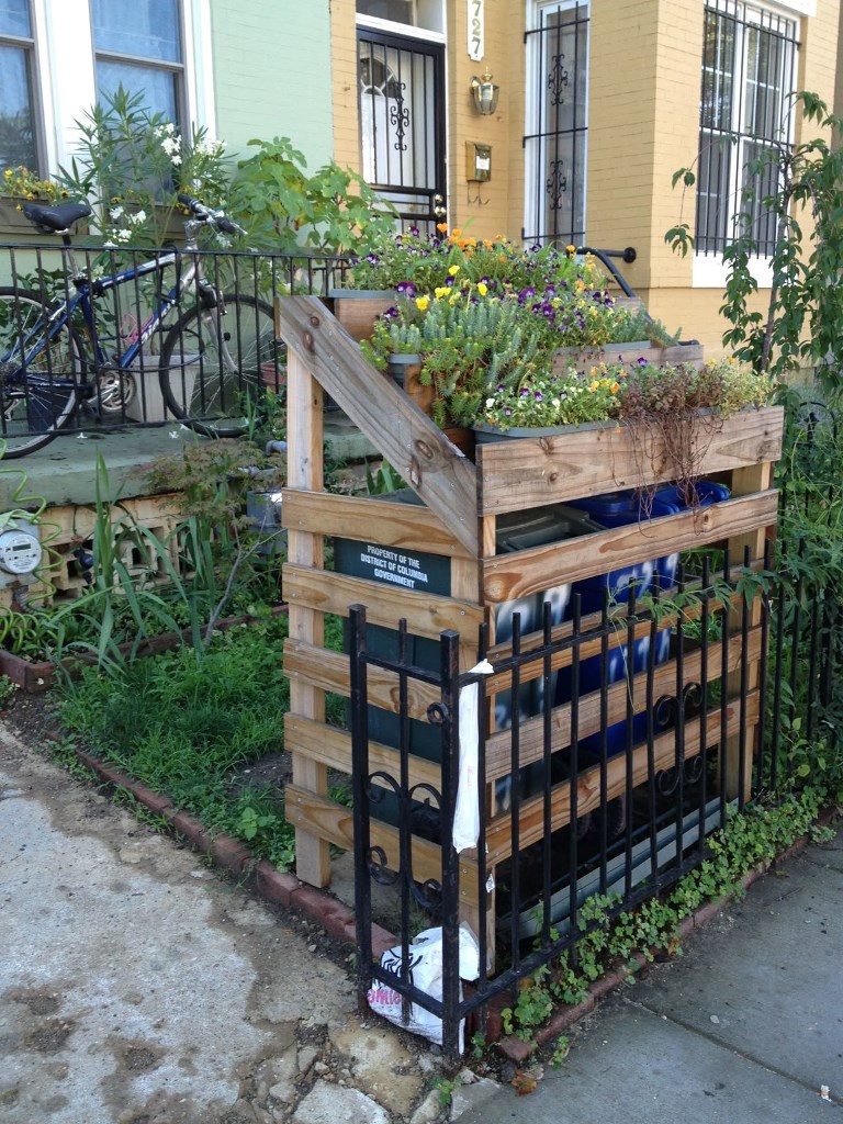 The trash cans are covered with a construction of pallet wood, and the tiered planters on top make up a cool look.