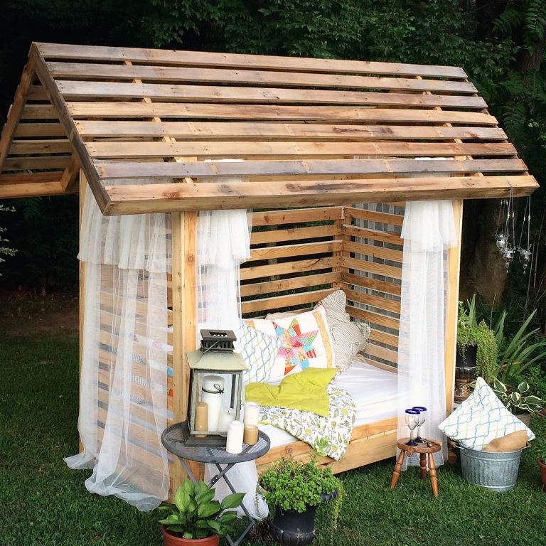 The little house-shaped pergola features a bed and some mosquito net curtains. Lanterns, greenery and wine glasses are the decor around.