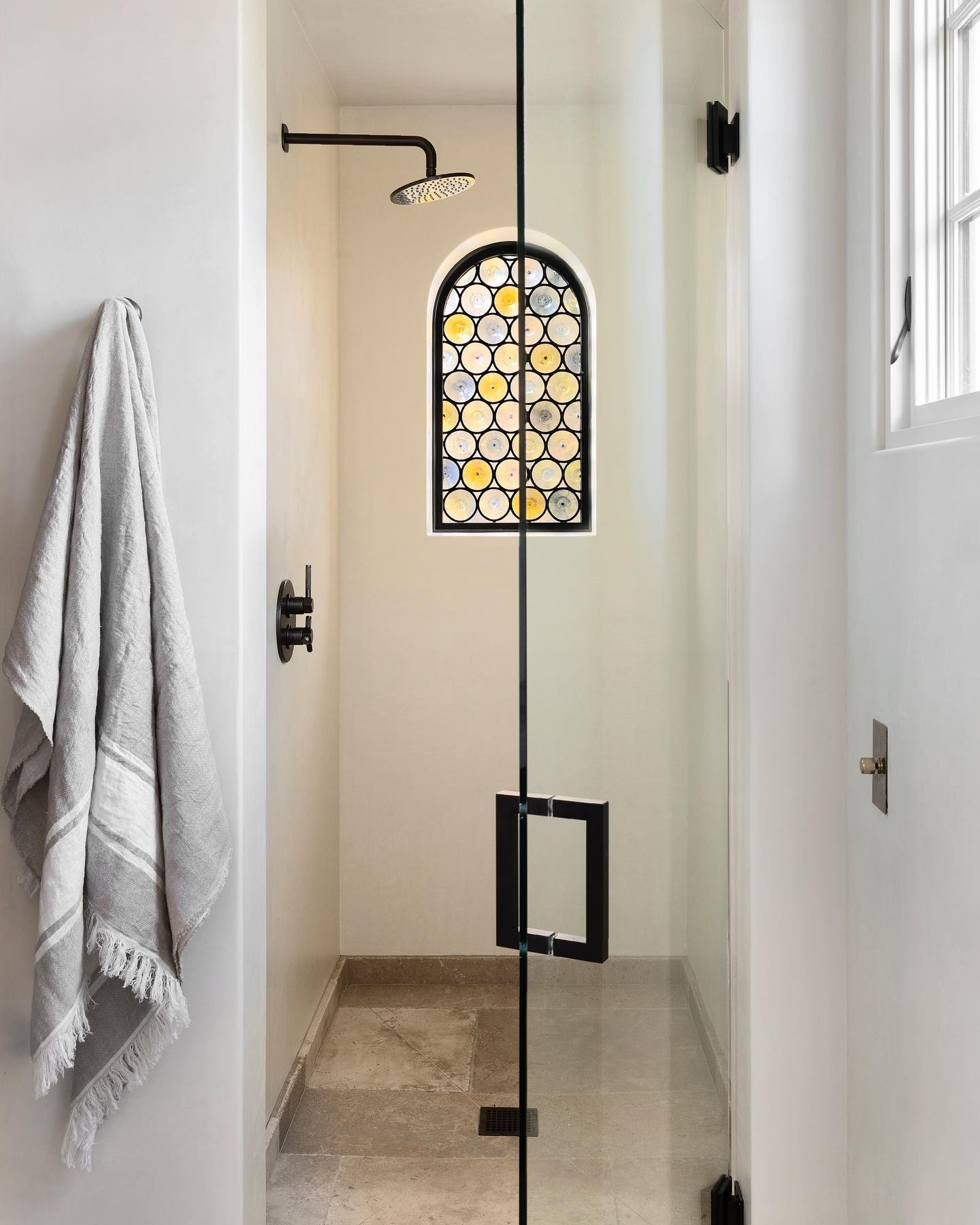 Moody with stained glass charm – A rondel window with warm tones gives this sleek black-and-white shower a historic twist. The mix of glass, stone, and curves feels rich yet restrained.

(@studiosurface).