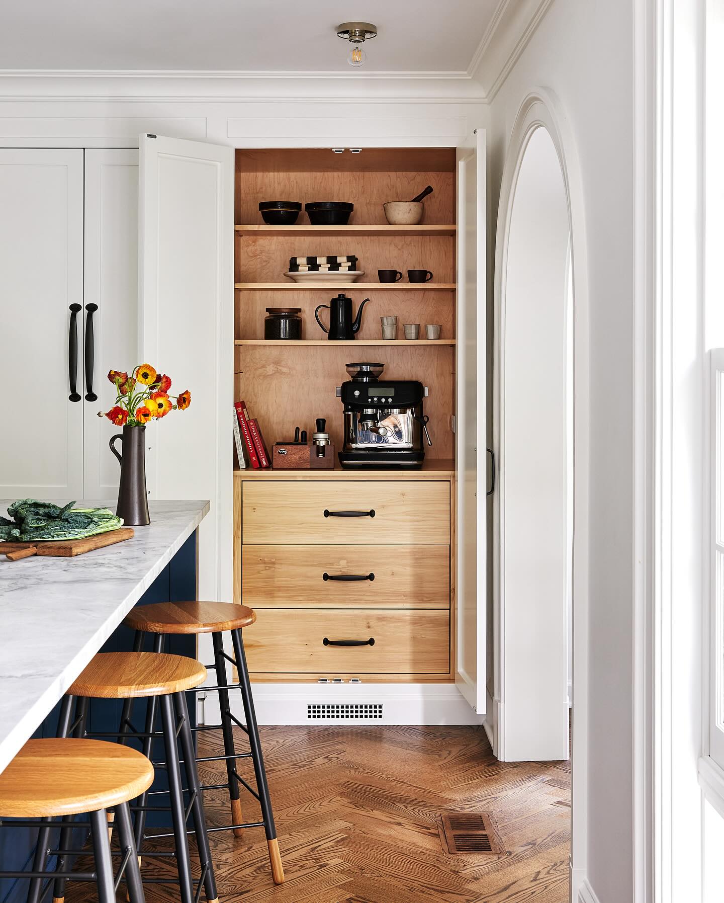This hidden kitchen pantry offers a stylish take on organization. Large wooden doors open to reveal deep shelving with neatly arranged dishware, a coffee machine, and small kitchen essentials. The black hardware and warm wood tones bring a cozy, sophisticated feel. (@thewhitearrow).