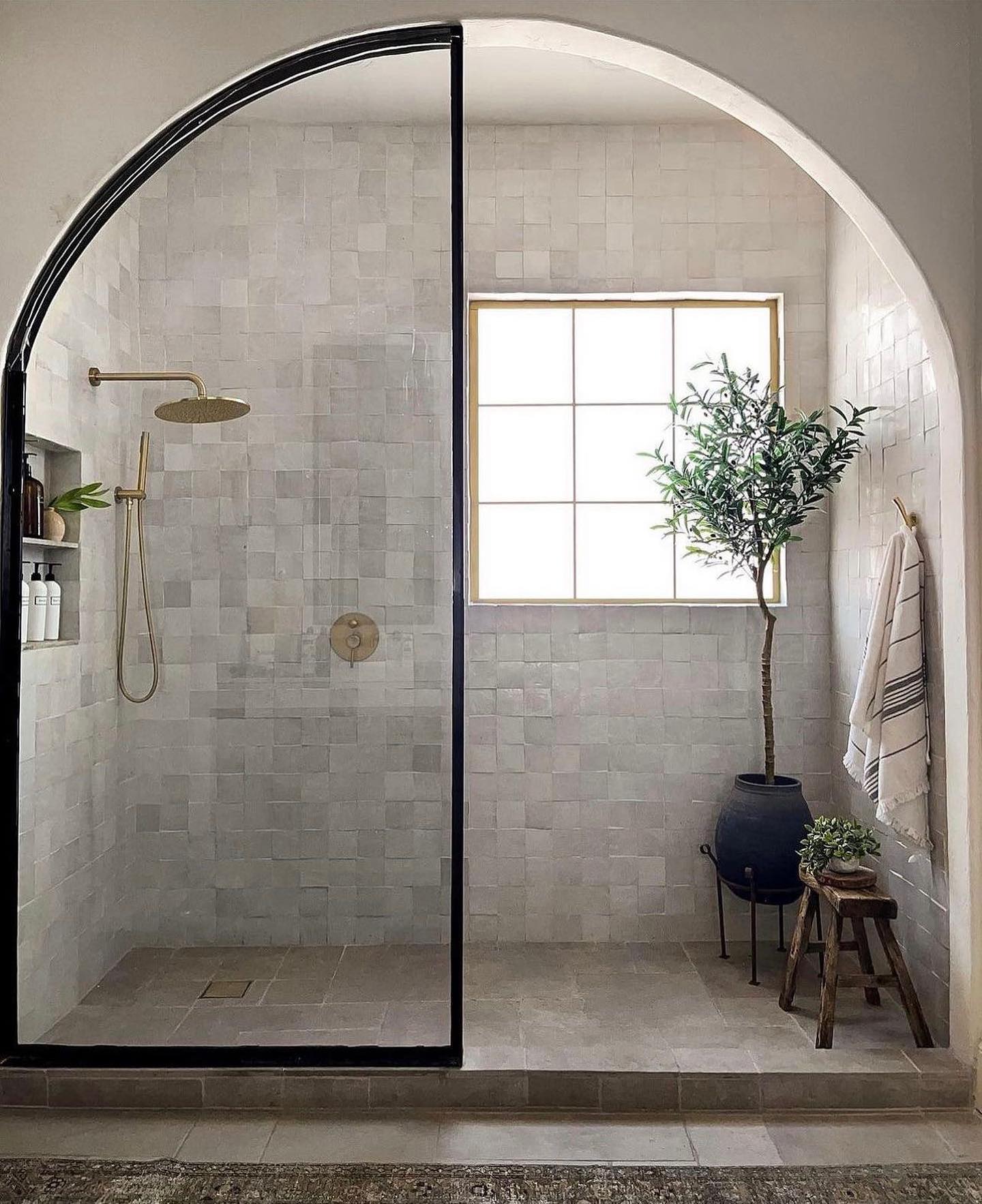 An arched shower entrance and a grid window create a spa-like retreat, complete with a little indoor tree and natural textures for a cozy organic feel.

(@puresaltinteriors).
