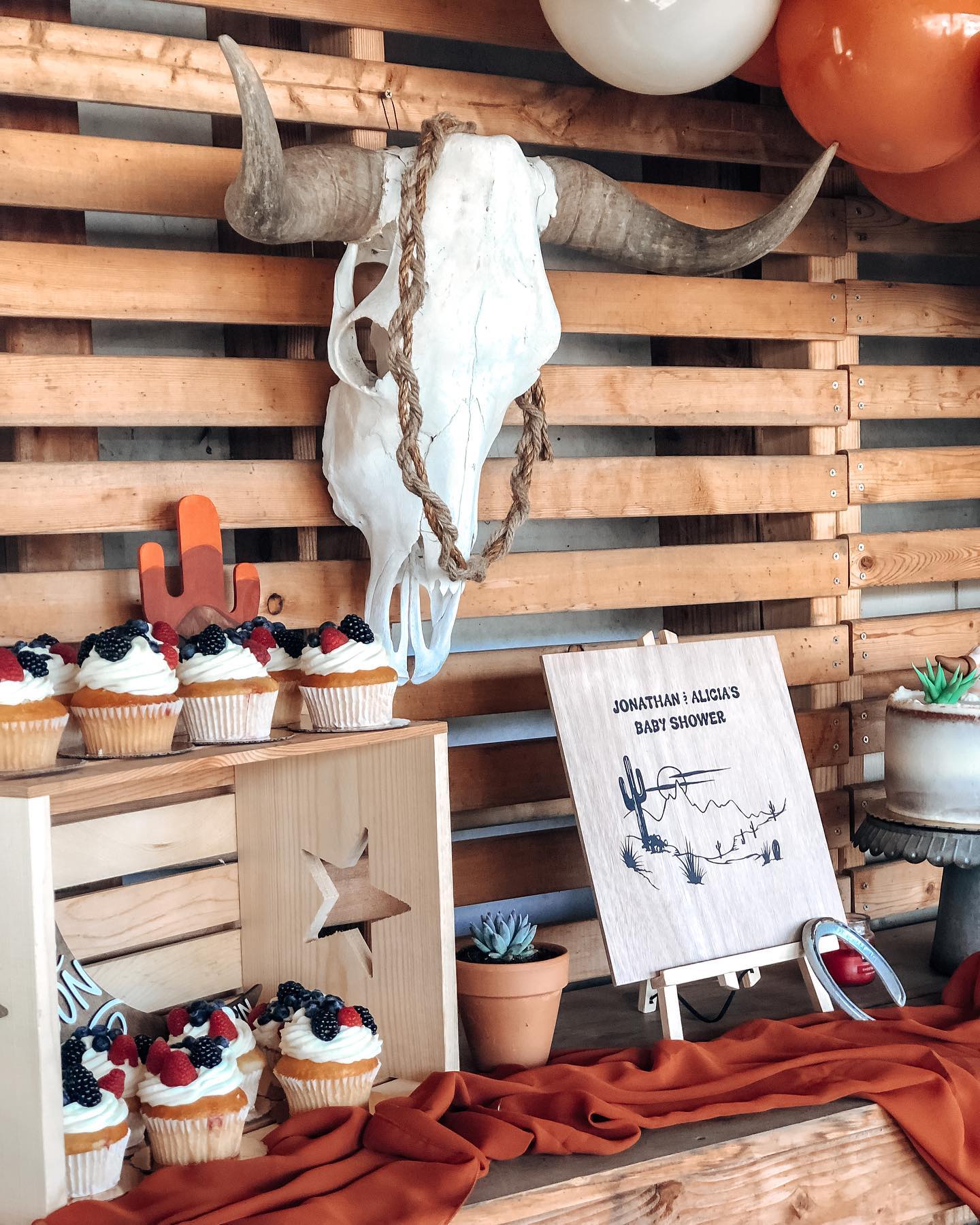 A rustic Western-themed baby shower display featuring a bold bull skull centerpiece, natural wood crates, cupcakes topped with fresh berries, and burnt orange fabric for a cozy, country feel.

(@ballinbridget).